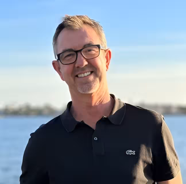 Dr. Michael Teichman, a Concord-based orthodontist, wearing glasses and a black polo shirt, smiling in front of a body of water.